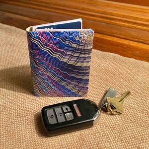 May include: A passport holder with a blue, purple, and gold abstract pattern. A car key with a silver key chain and a gold key is on a brown surface.