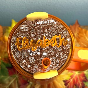 May include: A brown and gold tumbler lid with the name "Elizabeth" in gold lettering. The lid has a glittery finish and is decorated with fall-themed doodles and the words "Sweater Weather" and "Pumpkin Spice".