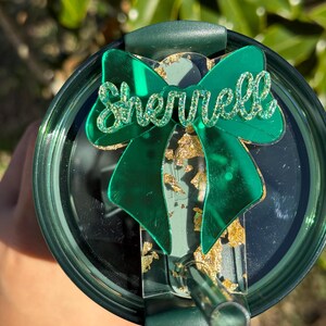 May include: Green tumbler with a green glitter bow and the name "Sherrell" on it. The tumbler has a gold glitter design.