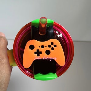 May include: A pink and green tumbler with a clear lid and a black and orange video game controller design. The lid has a straw hole and a black and green straw.