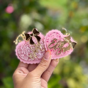 May include: Two pink heart-shaped keychains with gold accents and the name "Eliza" in gold script. Each keychain has a bow and is filled with glitter. The keychains are held up against a blurred green background.
