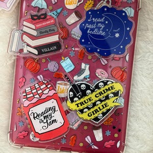 May include: A clear phone case with a burgundy background, decorated with various stickers. Stickers include a stack of books, pumpkins, a coffee cup, and the phrases "Reading is my Jam" and "True Crime Girlie."
