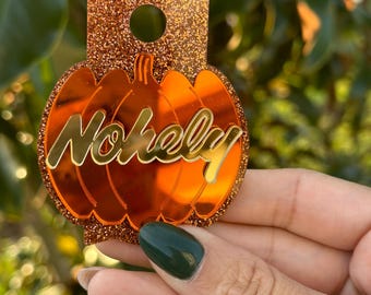 May include: A pumpkin-shaped acrylic ornament featuring a reflective orange surface with the name "Nohely" in gold script lettering. The pumpkin is layered over a rectangular backing piece covered in a dark orange glitter finish with a circular hole.