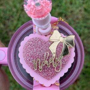 May include: A pink tumbler with a decorative lid and a glittery heart-shaped embellishment. The heart has the word "Nobely" in gold script and a gold bow. The lid has a pink floral design.
