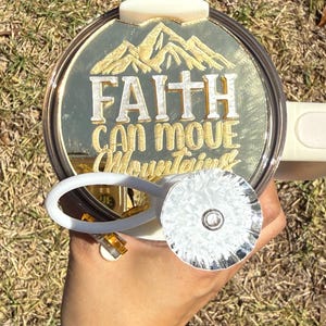 Puede incluir: Una tapa de vaso blanca con un diseño dorado que dice "La fe puede mover montañas". La tapa tiene un portapajitas plateado.