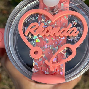 May include: A pink tumbler with a clear lid and a pink heart-shaped charm featuring the name "Rhonda" and a stethoscope design. The charm is covered in iridescent glitter.