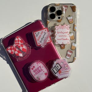 May include: A pink phone case with a clear back and various stickers. The stickers include a bow, a book stack, a coffee cup, and a heart with the text "Girls just wanna read books".