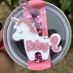 Peut inclure: Un gobelet transparent avec un couvercle et une paille roses, orné d'un motif de licorne blanche avec des accents de paillettes roses. Le nom "Riley" est écrit en paillettes roses. Le gobelet est conçu pour les boissons froides.