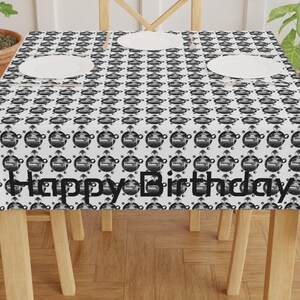 May include: A white tablecloth with a repeating black and white pattern of a birthday cake design. The words "Happy Birthday" are printed in black across the front of the tablecloth. Three white plates are on the table.
