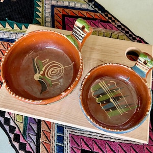 May include: Two handmade, terracotta-colored serving bowls with handles, resting on a light wooden board. The bowls feature painted interior designs in green, white, and blue. The handles are painted with green and blue accents. The bowls are set against a colorful, patterned textile.