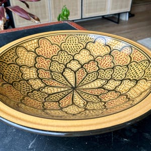 May include: A decorative ceramic bowl with a floral design. The bowl has a beige exterior and an intricate interior pattern in yellow, brown, and black. The bowl is placed on a dark surface, and is round in shape.