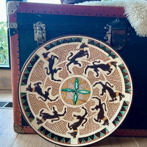 May include: A round, decorative plate with a brown border and a central design featuring a cross shape. The plate's surface is adorned with stylized black animal figures and geometric patterns in brown, beige, and green.