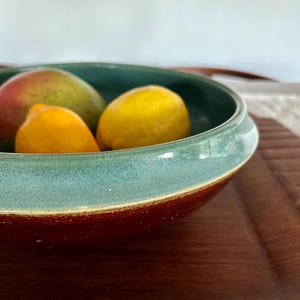 Puede incluir: Un cuenco de cerámica con borde turquesa y base marrón rojizo contiene un mango, un limón y una pequeña fruta naranja. El cuenco descansa sobre una superficie de madera, creando una naturaleza muerta natural y colorida.