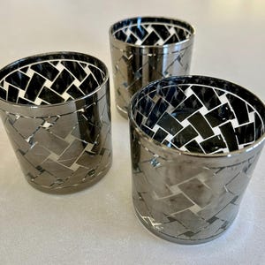 Puede incluir: Tres portavelas plateados. Cada soporte cilíndrico tiene un patrón tejido de baldosas plateadas y negras en el exterior. El interior de cada soporte es blanco y negro. Los soportes están dispuestos sobre una superficie de color claro.