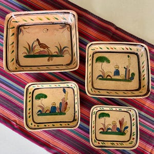 Może przedstawiać: Zestaw czterech kwadratowych naczyń ceramicznych z zaokrąglonymi rogami, o ręcznie malowanych powierzchniach w kolorze beżowym z brązowymi, zielonymi i niebieskimi motywami. Największe naczynie przedstawia ptaka, a trzy mniejsze postać przy drzewie i kaktusie.