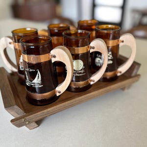 May include: Set of six amber-colored glass mugs with wooden handles and copper bands, resting on a wooden tray. Each mug features a white graphic, including a sailboat and golf ball. The tray is a dark brown color.