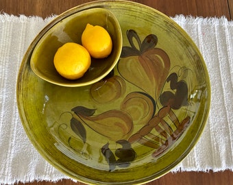 Puede incluir: Un plato de servir de cerámica verde con un diseño floral marrón y negro. El plato tiene un tazón más pequeño en el interior para servir aperitivos o salsas. Tres limones amarillos están en el tazón.
