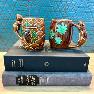 May include: Two ceramic mugs with squirrel handles. One mug is brown with a pineapple design and a pinecone. The other mug is brown with green leaf accents. Both mugs are on top of two books, one titled "The Nomad Cookbook".
