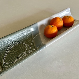 May include: A rectangular ceramic serving dish with a white and gray color scheme. The dish has a textured, speckled surface on one side and holds three bright orange tangerines. White abstract lines decorate the surface.
