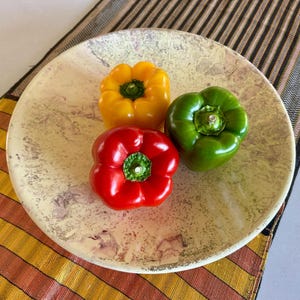 Puede incluir: Un plato de cerámica beige con tres pimientos: uno amarillo, uno rojo y uno verde. Los pimientos están colocados en un plato con una superficie moteada. El plato descansa sobre un camino de mesa a rayas con tonos naranja, amarillo y marrón.