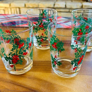 Könnte beinhalten: Vier klare Trinkgläser mit einem festlichen Stechpalmen- und Beeren-Design. Die Gläser sind mit grünen Stechpalmenblättern und roten Beeren verziert, ideal für Feiern. Sie sind auf einem Holztablett angeordnet.