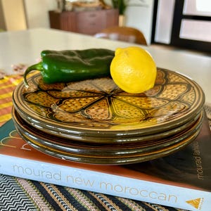 May include: A stack of three decorative plates with a brown and yellow floral design. A green pepper and a yellow lemon rest on top of the plates. The plates are stacked on a book with the title "mourad new moroccan".