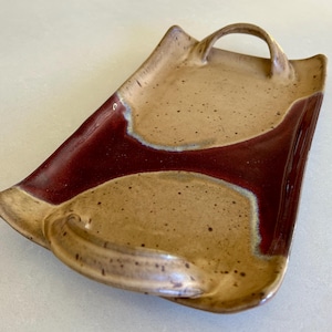May include: A rectangular ceramic serving tray featuring a speckled tan finish with a deep red glazed center section. The tray includes two arched handles on opposite ends, with curved edges and a smooth, glossy surface texture throughout the piece.