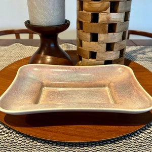 Puede incluir: Un plato rectangular de cerámica con un esmalte rosa claro y blanco. El plato tiene un borde ligeramente elevado y un acabado liso y brillante.