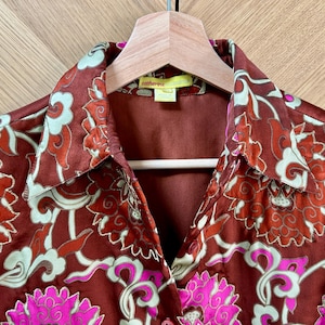 Può includere: Una camicia di raso marrone con una stampa floreale in tonalità di rosa, verde e bianco. La camicia ha un colletto e una chiusura con bottoni sul davanti. L'etichetta sulla camicia recita "Catherine".