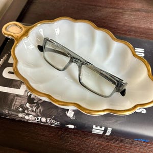 May include: A white, leaf-shaped dish with a gold rim and handle holds a pair of eyeglasses. The dish sits atop a book with black and white imagery and text. The dish is a decorative item.