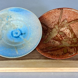 Può includere: Due ciotole in ceramica, una con una glassa blu e bianca e l'altra con una glassa marrone e arancione. Entrambe le ciotole hanno un design astratto unico.