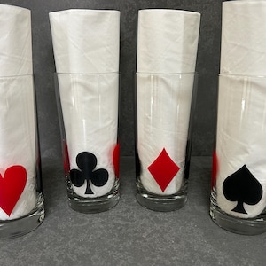 Op de afbeelding: Vier heldere drinkglazen met speelkaartsymbolen: een rode harten, een zwarte klaver, een rode ruiten en een zwarte schoppen. Elk glas bevat een gevouwen witte servet. De glazen staan op een grijze ondergrond.