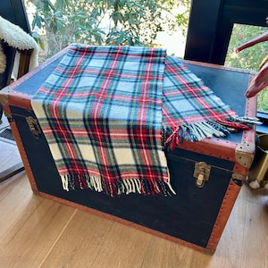 Puede incluir: Una bufanda a cuadros rojos, verdes, blancos y azules está sobre un baúl vintage. La bufanda tiene bordes con flecos y un patrón de tartán clásico. El baúl es negro con ribetes marrones y herrajes metálicos.