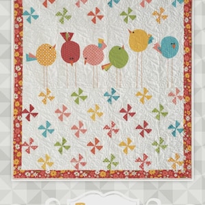 May include: A white quilt with a red floral border features six colorful bird appliqués on thin stems. Pinwheel designs in various colors are scattered across the quilt's surface. The word "Twitter" is displayed in a gold font.
