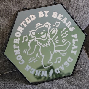 May include: Hexagon-shaped mirror with a black frame. The mirror features a white graphic of a dancing bear with musical notes and the text "WHEN CONFRONTED BY BEARS PLAY DEAD". The mirror is displayed on a gray textured surface.
