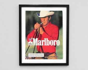 May include: Framed vintage Marlboro advertisement showing a cowboy in a red shirt and white hat lighting a cigarette. The Marlboro logo is displayed. The image is in a black frame.