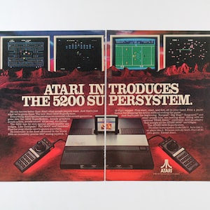 May include: A vintage Atari 5200 Supersystem video game console advertisement. The ad features a red, white, and blue color scheme and shows the console with two controllers. The text "ATARI INTRODUCES THE 5200 SUPERSYSTEM." is displayed in large, bold letters.