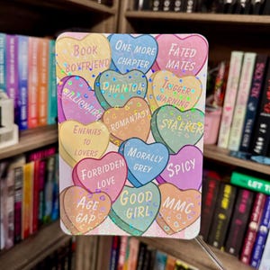 May include: A rectangular sticker featuring colorful heart-shaped candies with text like "Book Boyfriend," "Fated Mates," and "Trigger Warning." The hearts have a holographic, iridescent finish. The background shows bookshelves filled with books.