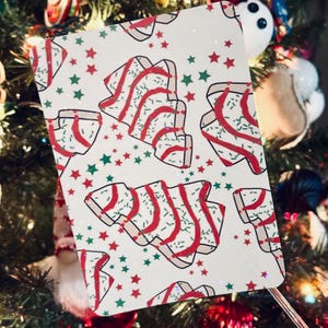 Puede incluir: Una tarjeta rectangular con un diseño de árbol de Navidad. La tarjeta presenta un fondo blanco con estrellas rojas y verdes, e ilustraciones de árboles de Navidad con glaseado rojo y chispas. La tarjeta se muestra frente a un árbol de Navidad decorado.