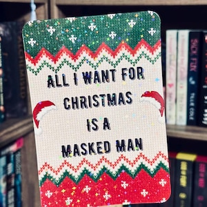 May include: A rectangular Christmas card with the text "ALL I WANT FOR CHRISTMAS IS A MASKED MAN". The card features a festive sweater design in red, green, and white, with Santa hats on either side of the text.