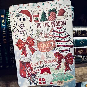 Puede incluir: Una pegatina rectangular con temática navideña con varias ilustraciones festivas. El diseño incluye un fantasma con un gorro de Papá Noel, árboles de Navidad, lazos y el texto "'Tis the Season" y "Let It Snow somewhere else". La pegatina está en una estantería.
