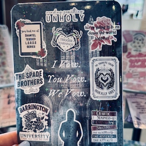 May include: A dark, rectangular notebook covered in stickers. Stickers include text like "Unholy", "Trigger Warnings", and "The Spade Brothers". Other stickers feature roses, skulls, and the words "I Vow".