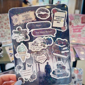 May include: A dark, glittery sticker sheet featuring various gothic-themed stickers. Designs include a gun, lips, books, a skull, and text that reads "Zade Meadows stole my morals and my soul." Other text includes "Trigger Warnings" and "Yes, Little Mouse?"