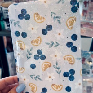 May include: A rectangular, glittery, light blue item with a pattern of blueberries, lemon slices, white flowers, and green leaves. The design is repeated across the surface. The item has rounded corners.