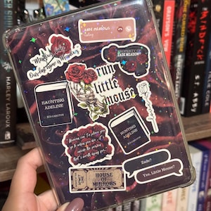 以下が含まれることがあります： バラ、本のタイトル、「Run Little Mouse」や「House of Mirrors」などのフレーズが描かれたステッカーで飾られた透明な保護ケース。ケースは本の背景に配置され、文学的なテーマを作り出しています。