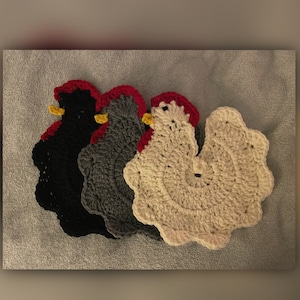 May include: Three crocheted chicken pot holders in black, gray, and white. The black pot holder has a red comb and wattle, the gray pot holder has a red comb and wattle, and the white pot holder has a red comb and wattle.
