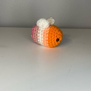 May include: A crocheted bee toy with pink, white, and orange stripes. The bee has black eyes and white wings.