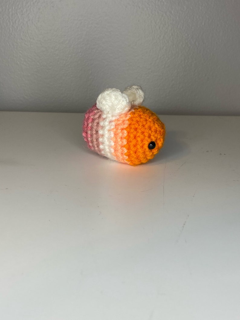May include: A crocheted bee toy with pink, white, and orange stripes. The bee has black eyes and white wings.