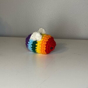 May include: A crocheted rainbow bee with white wings. The bee has a black eye and is made with a variety of colorful yarn.