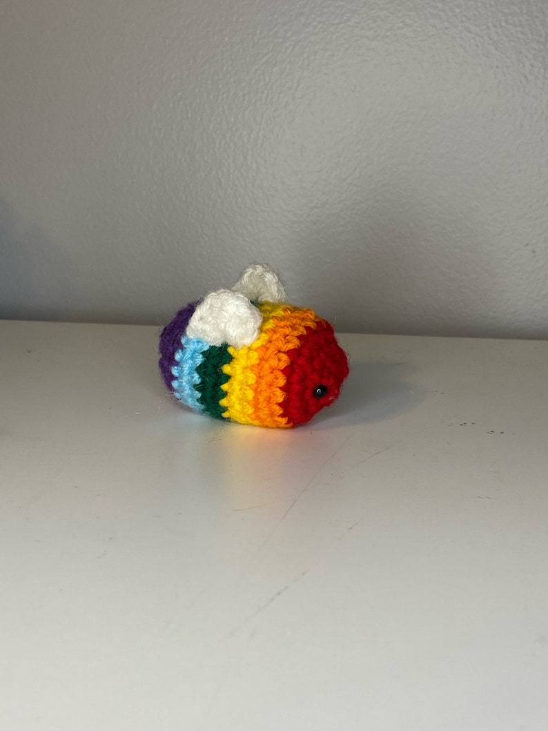 May include: A crocheted rainbow bee with white wings. The bee has a black eye and is made with a variety of colorful yarn.